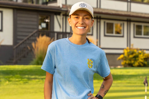 A 5'1" Woman in a small light blue shirt and cap standing on a golf course with a modern house in the background