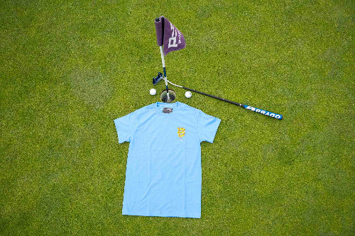 A light blue short-sleeve polo shirt with a small yellow crest on the left chest, laid flat on a green grass surface. A golf flag, putter, golf ball, and golf club are arranged above it as styling props.