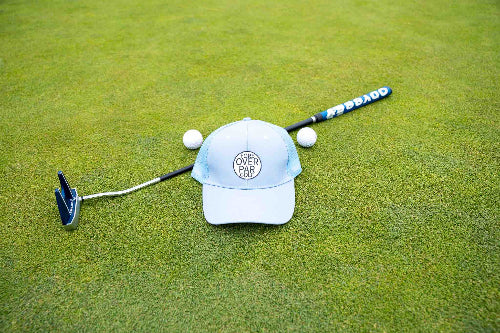 Light Blue Pony Tail Club hat by Fore Over Par shown on a golf green with a putter and two golf balls behind it. 