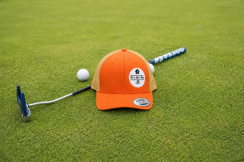 An orange and tan baseball cap with a 'Fore Over Par' logo, styled on a green grass surface with a golf putter, golf ball, and golf club.