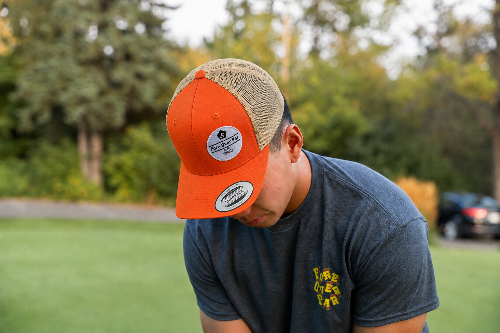 A 5'10" man outdoors wearing a two-tone baseball cap with a bright orange front and brim and a beige mesh back with embroidered "Fore Over Par", paired with a dark gray t-shirt with a colourful "Fore Over Par" Logo