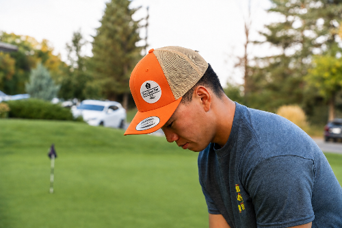 A 5'10"man on a golf course wearing a tan and orange trucker hat with a visible 'Fore Over Par' brand patch, looking down toward a golf hole flag.