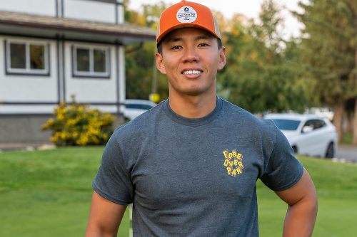 A 5'10" man wearing a dark gray t-shirt with a colourful 'Fore Over Par' Logo, black pants, and an orange cap with embroidered 'Fore Over Par' Logo stands on a golf course holding a golf club.