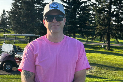 A man wearing a bright pink t-shirt, a white cap , and sunglasses stands on a golf course holding a golf club. A golf cart is visible in the background on the left, with tall evergreen trees and a blue sky behind him.