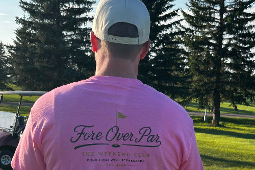A man viewed from behind wearing a pink t-shirt with 'Fore Over Par' printed on the back, standing on a grassy golf course with trees in the background.