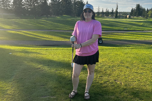A 5'5" woman stands on a golf course holding a golf club, wearing a pink t-shirt, black skirt, and a cap, with trees and a blue sky in the background.