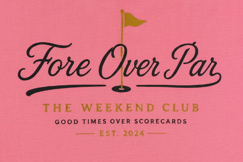 A close-up of a pink t-shirt with printed text reading 'Fore Over Par' and 'THE WEEKEND CLUB' plus smaller text 'GOOD TIMES OVER SCORECARDS' and 'EST. 2024', alongside a small gold flag graphic.