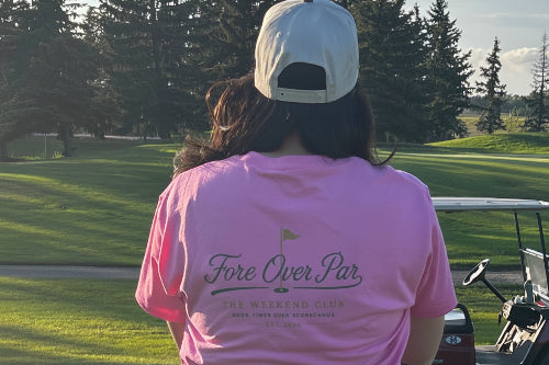 A woman standing on a golf course, viewed from behind, wearing a pink t-shirt with 'Fore Over Par' printed on the back, a black skirt, and a baseball cap.