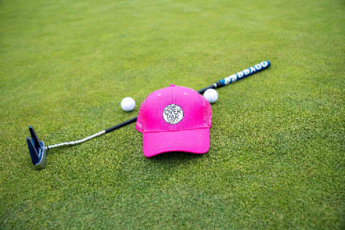 Pink Pony Tail Club by Fore Over Par shown on a golf green with a putter and two golf balls behind it. 