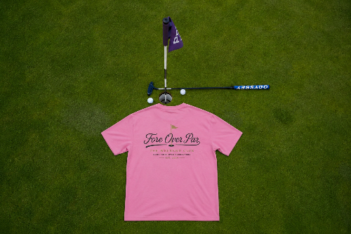 A pink short-sleeve t-shirt laid flat on a green grass background, with golf-themed props above it including a flagstick, putter, and golf balls. The shirt has black script text reading 'Fore Over Par' and smaller text below.
