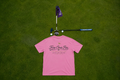 A pink short-sleeve t-shirt laid flat on a green grass background, with golf-themed props above it including a flagstick, putter, and golf balls. The shirt has black script text reading 'Fore Over Par' and smaller text below.