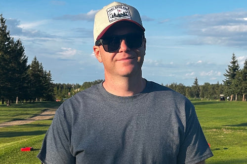A man wearing a gray t-shirt, black sunglasses, and a beige baseball cap with a red brim stands on a golf course. The cap features a logo that reads ' Fore Over Par'.