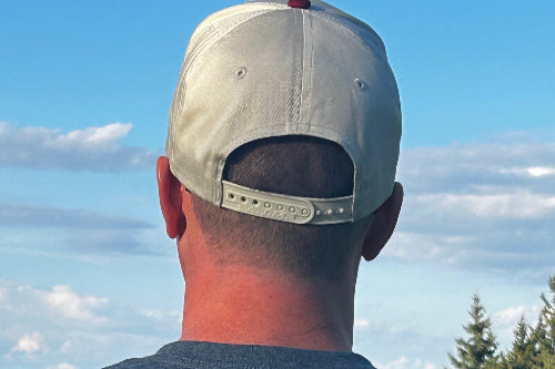 Rear view of a man wearing a light beige baseball cap with an adjustable strap and a dark gray t-shirt, set against a blue sky with clouds and trees.