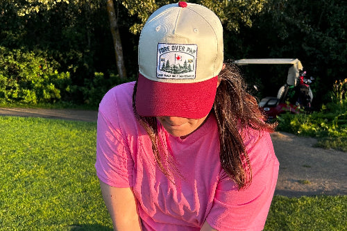 A person outdoors on a grassy area wearing a beige and red 'Fore Over Par' baseball cap, a pink t-shirt, and black shorts, looking down while holding a golf club.