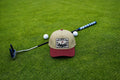 A beige and burgundy baseball cap with a 'Fore Over Par' logo, styled on a green grass background with golf props including a putter, golf balls, and a golf club.