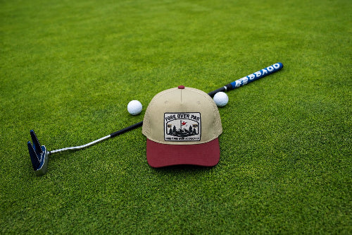 A beige and burgundy baseball cap with a 'Fore Over Par' logo, styled on a green grass background with golf props including a putter, golf balls, and a golf club.