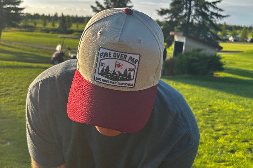 A man on a golf course wearing a light gray and maroon baseball cap with a patch that reads 'FORE OVER PAR'.