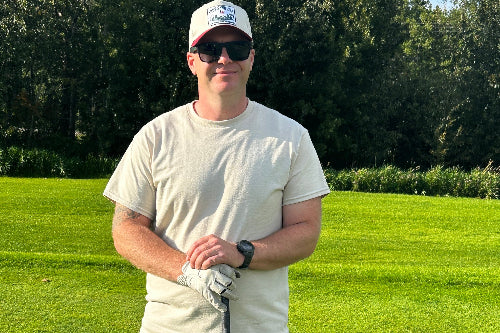 A man standing on a grassy golf course wearing a sand t-shirt, dark gray shorts, a white cap with a blue logo, sunglasses, and a white golf glove while holding a club.