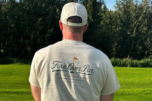 A man viewed from behind wearing a light sand t-shirt with a 'Fore Over Par' golf course graphic on the back, standing on a green grassy field with trees in the background.