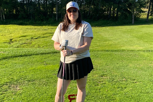 A woman stands on a grassy golf course holding a golf club, wearing a sand t-shirt, black pleated skirt, and a sand cap. 