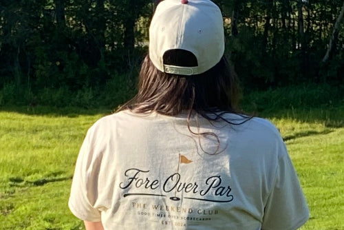 A person standing on a grassy golf course, viewed from behind, wearing a sand t-shirt with 'Fore Over Par' printed on the back, a black pleated skirt, and a sand baseball cap.