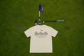 A sand-colored t-shirt with 'Fore Over Par' printed on the back, laid flat on a green grass background alongside a purple flag, golf club, and golf balls.