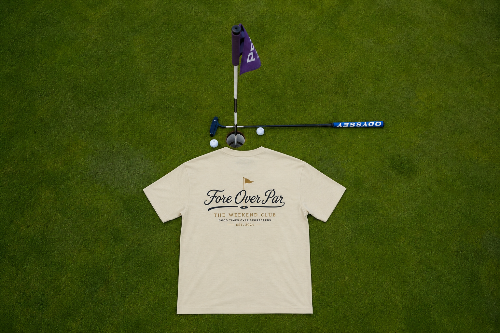 A sand-colored t-shirt with 'Fore Over Par' printed on the back, laid flat on a green grass background alongside a purple flag, golf club, and golf balls.