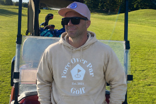 A 5'9" man wearing a sand coloured 'Fore Over Par Golf' hoodie, sunglasses, and a pink cap stands in front of a red golf cart on a sunny golf course.