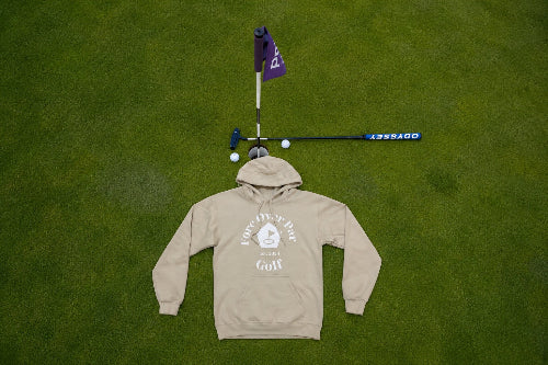 A sand pullover hoodie with a white 'Fore Over Par Golf' graphic is laid flat on a green grass surface, styled with a purple flag, golf club, and golf balls above it.