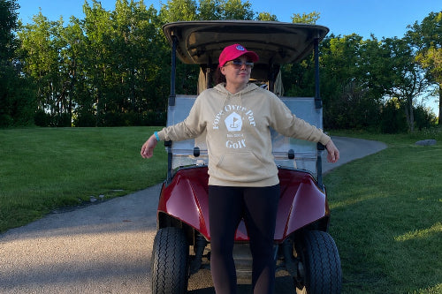 A 5'5" woman stands in front of a red and white golf cart on a paved path outdoors, wearing a sand coloured hoodie with a white 'Fore Over Par Golf' graphic, black leggings, and a pink cap. Trees and grass are visible in the background under a clear blue sky.