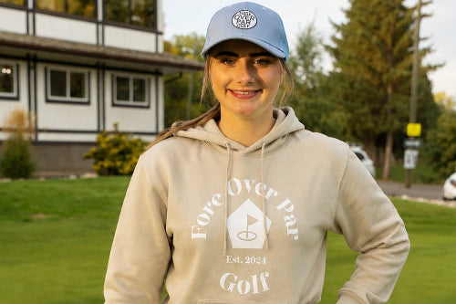 A 5'10" woman stands on a golf course wearing a sand coloured hoodie with a white 'Fore Over Par Golf' graphic, and a light blue cap.
