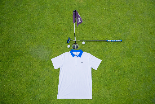 A white short-sleeve polo shirt with a blue collar is laid flat on a green grass surface, styled with golf props including a flagstick, putter, golf balls, and tees above it.