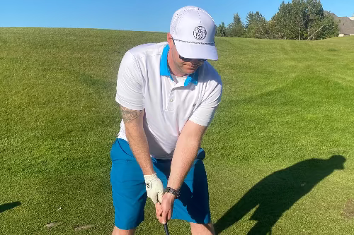 A 5'9" man wearing a white polo shirt with a blue collar, blue shorts, and a white cap is shown from behind on a golf course, holding a golf club. The background features a green grass field and a clear blue sky.