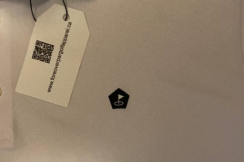 Close-up of a white fabric surface with a small black pentagonal Fore Over Par logo near the center and a hanging product tag on the upper left