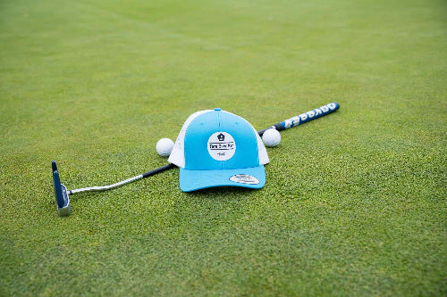 Turquoise & White OG Hat by Fore Over Par shown on golf green with a putter and two golf balls behind it.