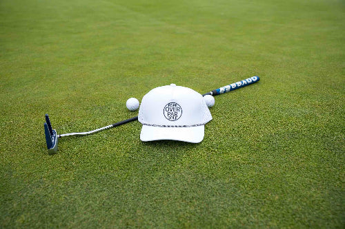White 19th Hole Rope Hat by Fore Over Par shown on a golf green with putter and two balls behind it. 